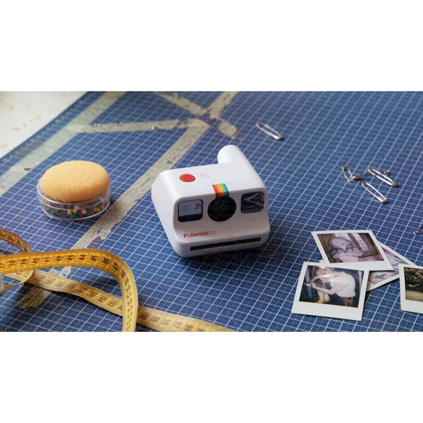 White camera on a blue cutting mat beside instant prints, a tape measure and a pincushion.